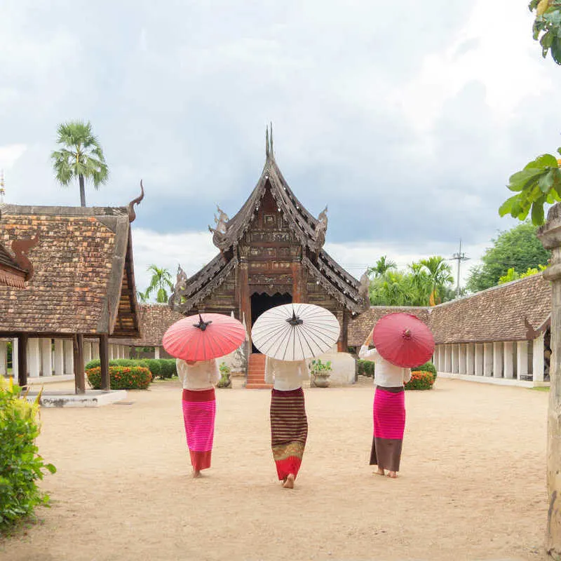 Thai Women Wearing Traditional Attire And Holding Up Traditional Umbrellas As They Walk Towards A Temple In Chiang Mai, Thailand, Southeast Asia