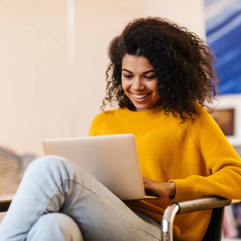 Young Smiling African American Woman Wearing A Yellow Pullover While Working From Her Computer In A Living Room Or Office Setting, Booking Flights Or Digital Nomad Concept