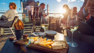 People eating cheese and drinking wine at rooftop restaurant at sunset time. Restaurant table served with cheese plate, bread and white vine full of visitors.