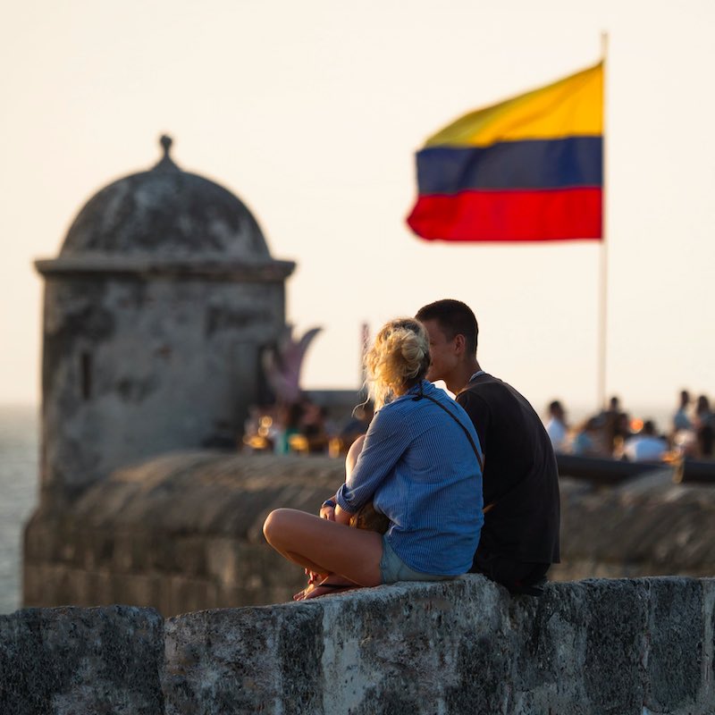 two travelers sitting on the city wall for sunset in Cartagena, the Colombian national flag waving in the background