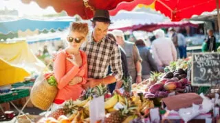 a young couple buying fruits and vegetables in a market on a sunny morning, the young woman carries a basket