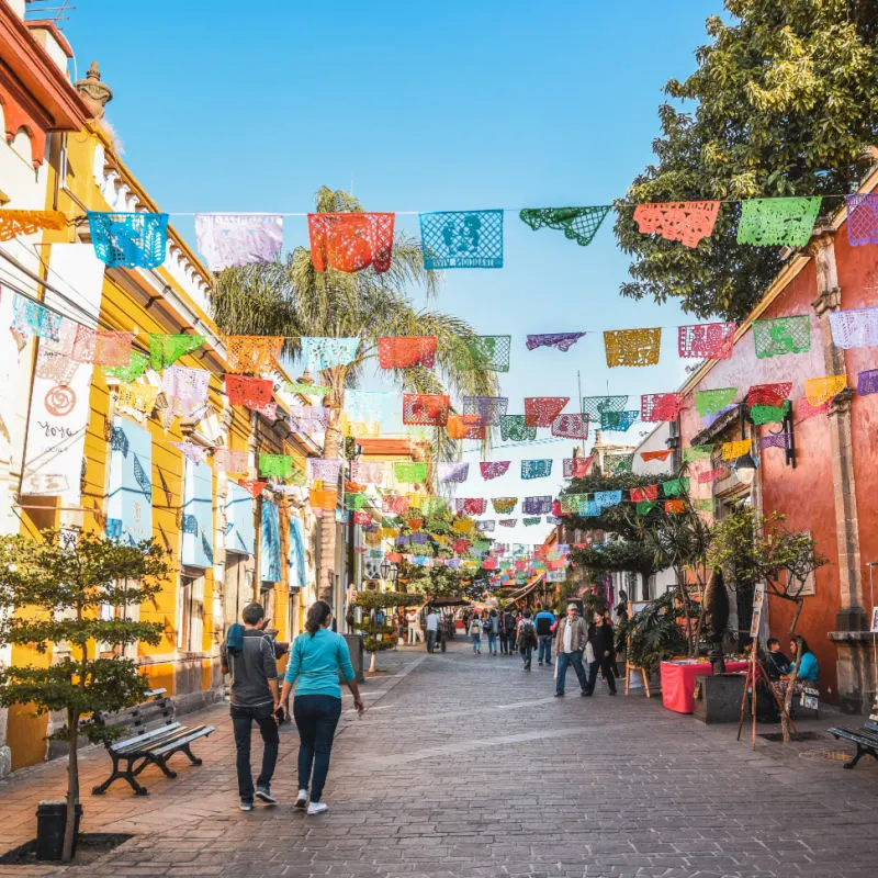 People walking through the colorful streets of Tlaquepaque, Mexico with papel picado overhead