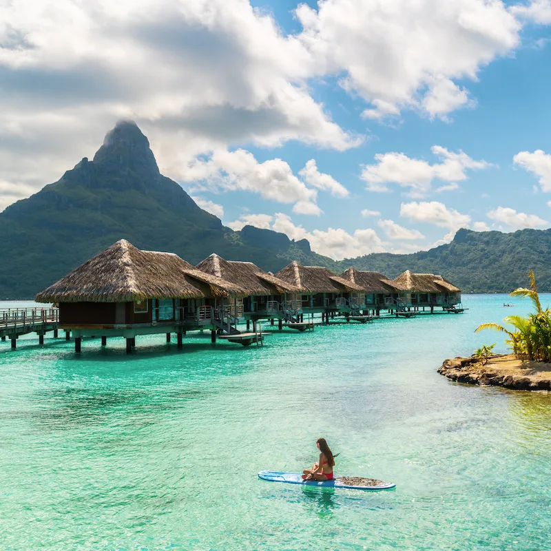 A woman rests on a paddleboard in front of a row of overwater bungalows in Bora Bora