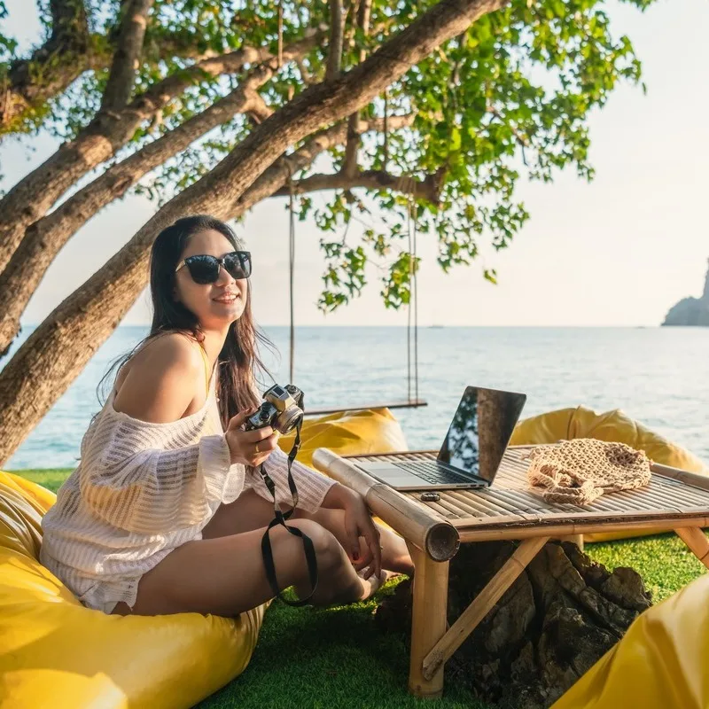Young Thai Woman Sitting Facing Her Computer As She Takes Photographs By The Beach In Phuket, Thailand, Southeast Asia